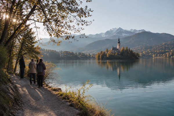 A small group on a lakeside path looking out across green water to a wooded island, Lake Bled, Bled