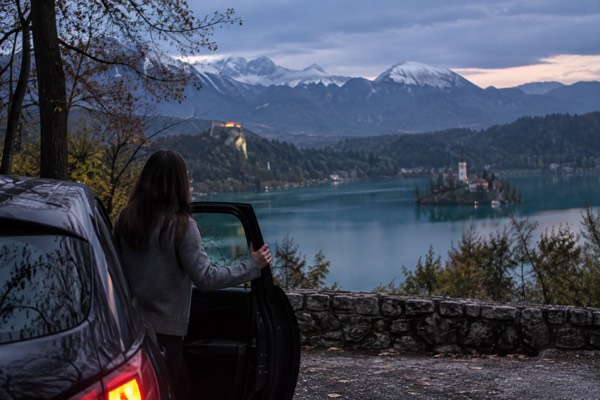 A private car pulled in at a lakeside viewpoint, its driver holding the door, green water and wooded slopes beyond, Lake Bled, Bled