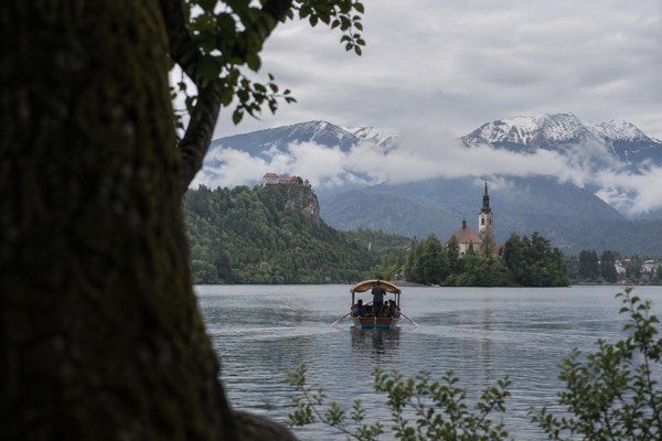 A small hilltop castle on a crag above a green lake, a flat-bottomed wooden boat crossing towards a wooded island below it, Lake Bled, Bled