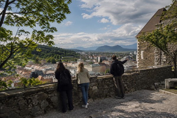 Two visitors and a guide on a castle rampart looking out over tiled rooftops towards distant hills, Ljubljana Castle, Ljubljana