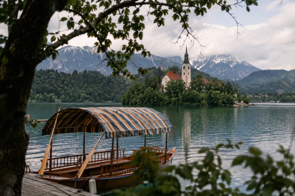 A wooden pletna boat with a canvas canopy resting on the shore of a lake beside a church-topped island