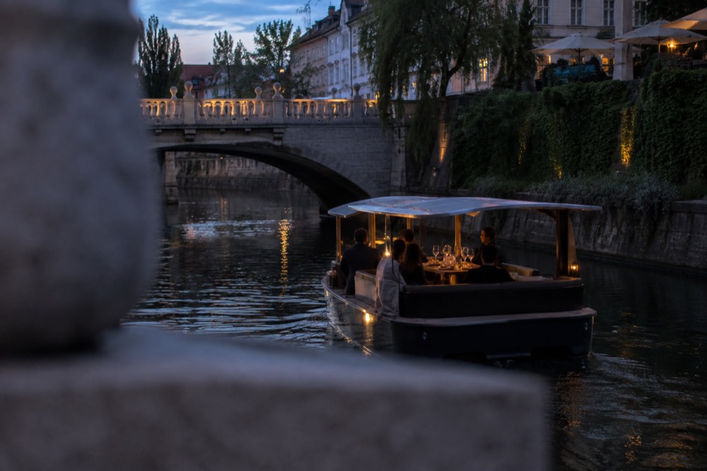A small electric boat on the Ljubljanica at dusk with wine glasses and appetizer plates arranged on a table