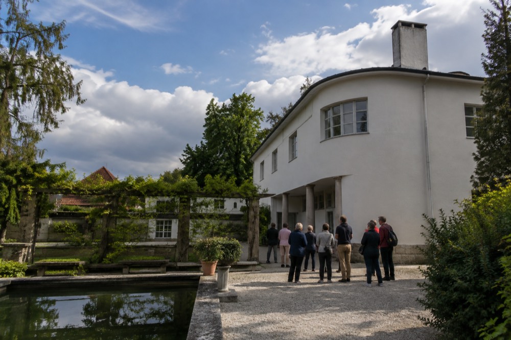 A group of visitors listening to a guide beside a white villa with a rounded corner