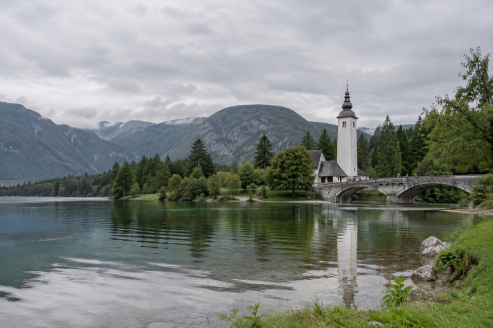A stone bridge over a river outflow with a small church with a tall tower rising above the water