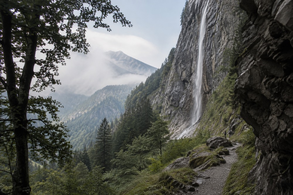 A tall thin waterfall falling down a grey rock face with a walking trail below it