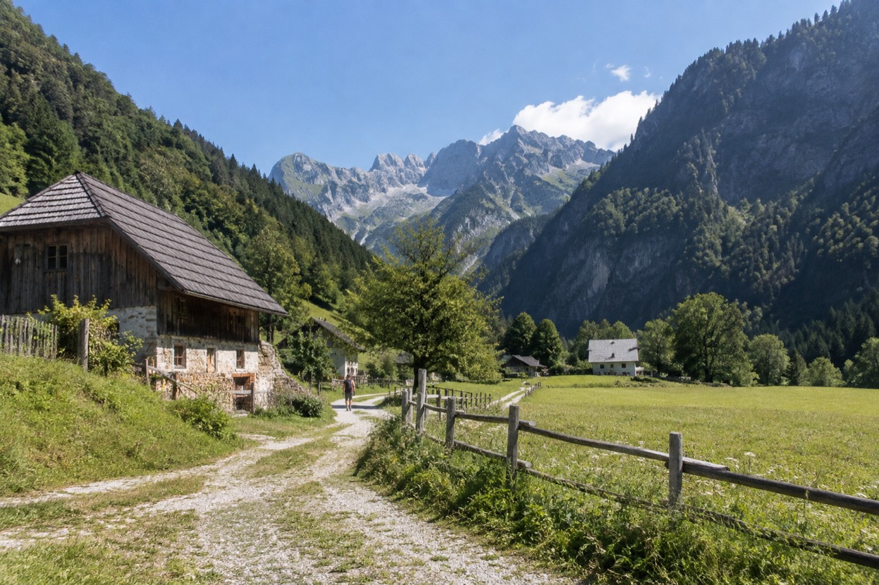 A narrow alpine valley with green meadows, wooden farmhouses and steep forested slopes rising to rocky peaks under a blue sky