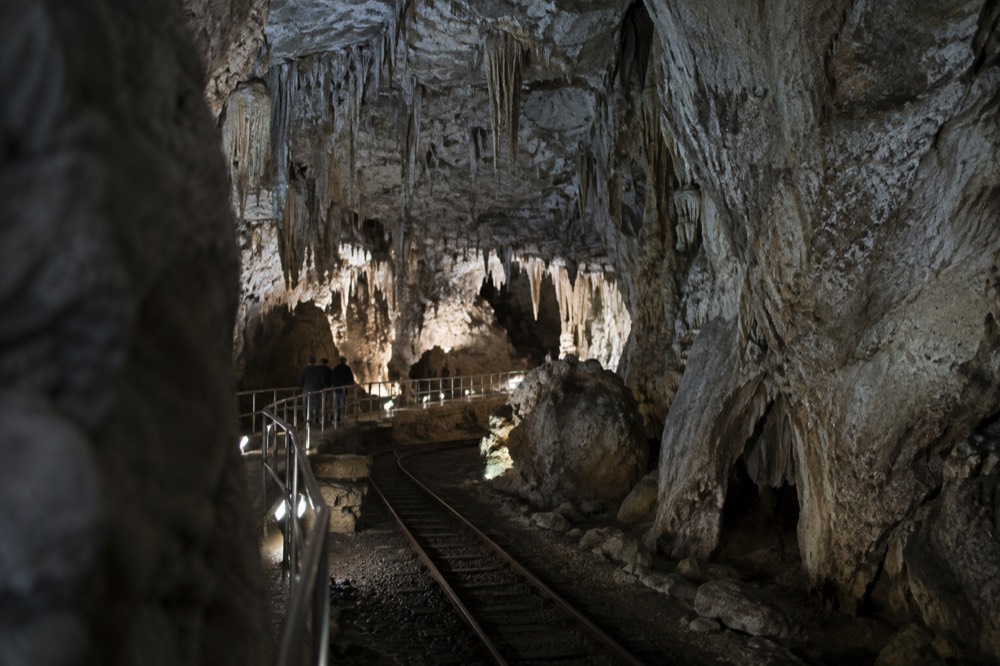 A cave chamber with stalactites lit by walkway lights above a rail line