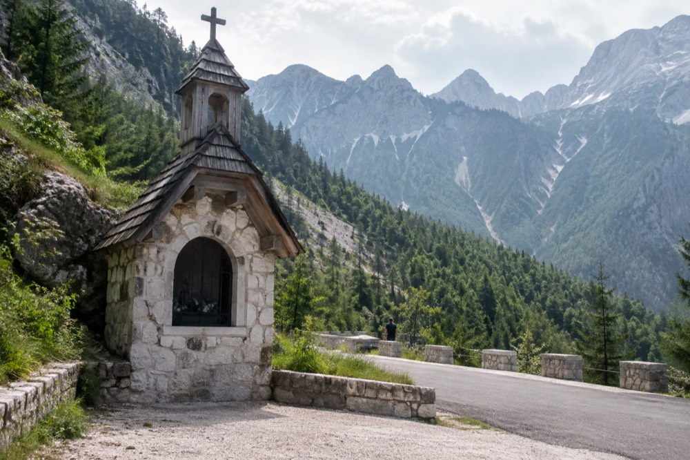 A stone roadside chapel beside a mountain road with steep green slopes rising behind it