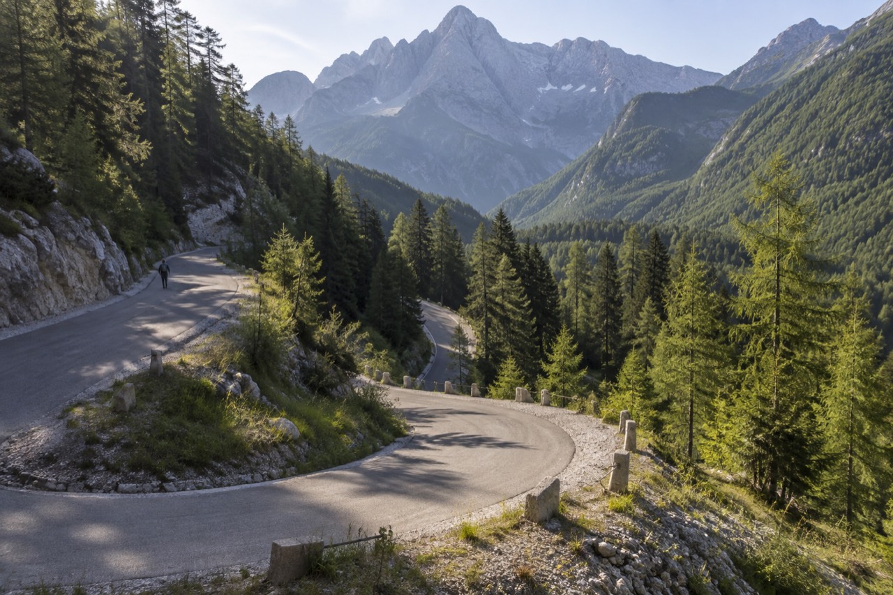 A winding mountain road with hairpin bends climbing through green alpine forest toward rocky peaks