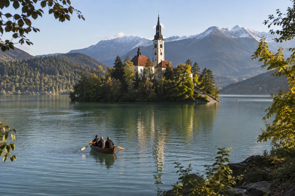 A wooden boat on a green lake approaching a small island with a church tower and wooded shores behind