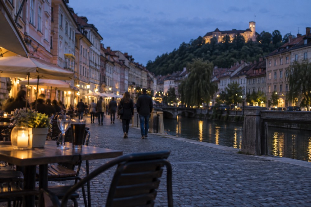 People strolling along a cobbled riverside street lined with outdoor tables in central Ljubljana