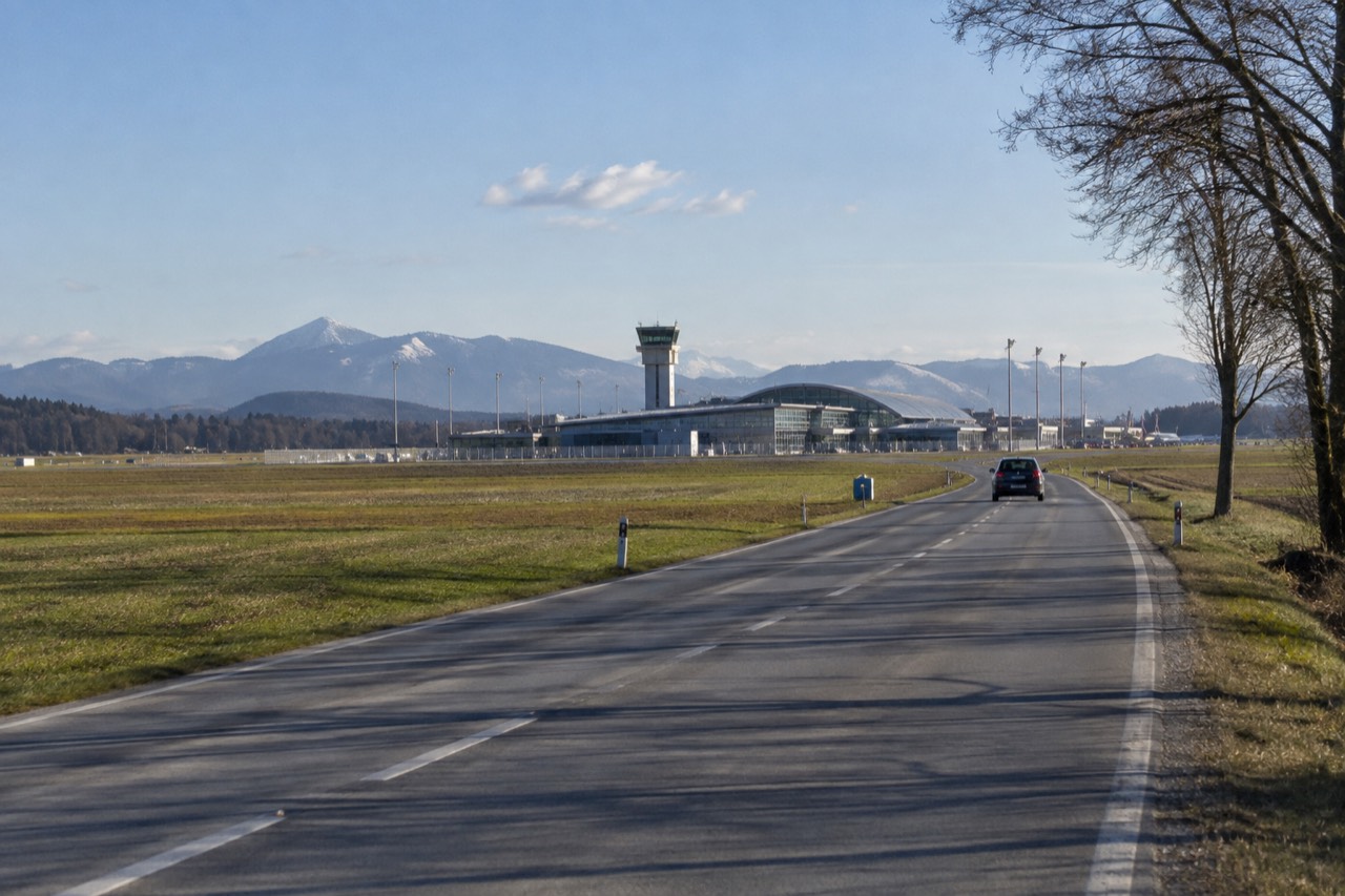A quiet road with green fields and distant hills leading toward a small airport terminal building under a blue sky.