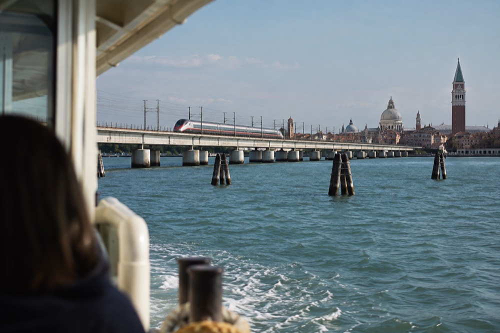 A high-speed train crossing a long bridge over a lagoon towards a city skyline